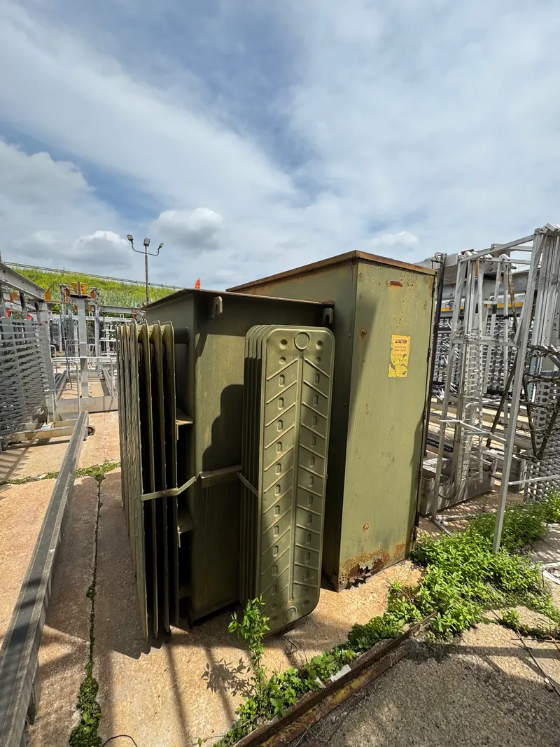 Older liquid-filled electrical transformer with radiator fins inside an industrial substation yard