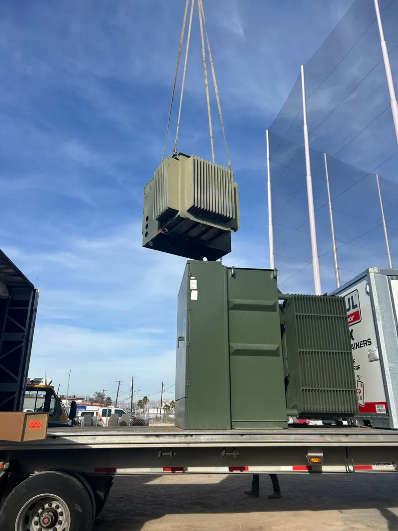 Industrial transformer suspended by lifting straps above a flatbed during crane loading
